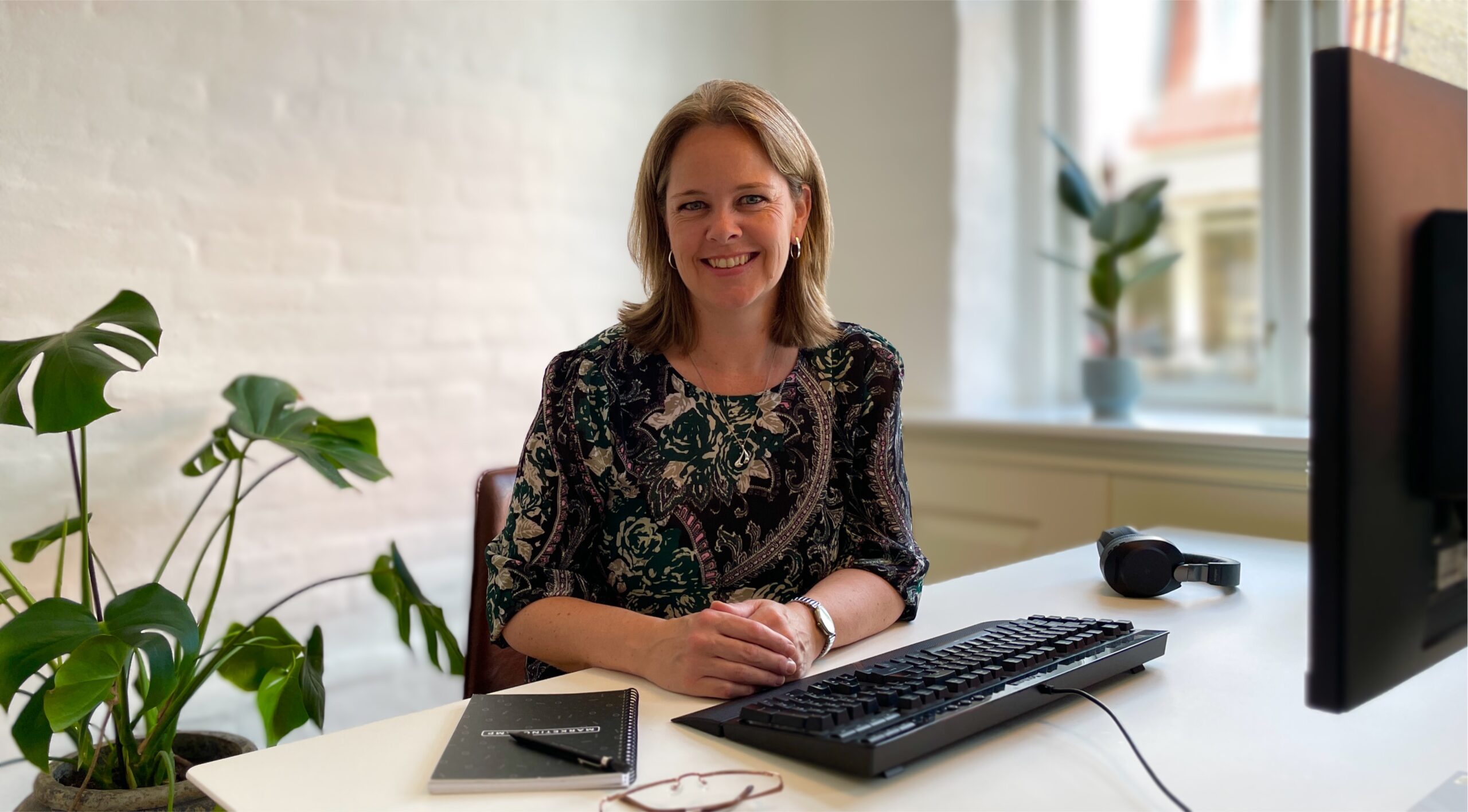 Smiling woman sitting at an office desk. Glasses, pen, paper and keyboard in front of her.