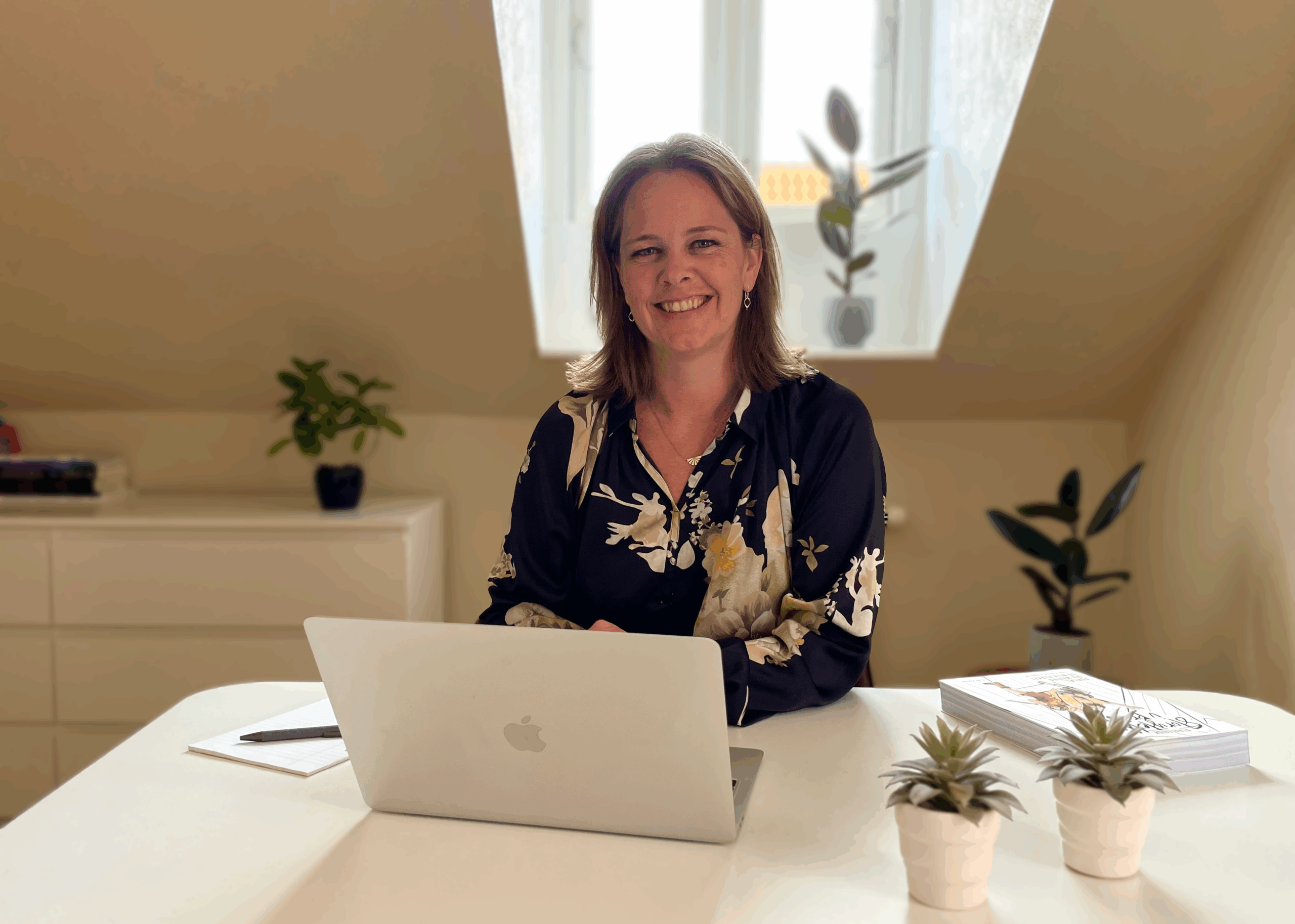 Smiling woman sitting at an office desk. Glasses, pen, paper and keyboard in front of her.